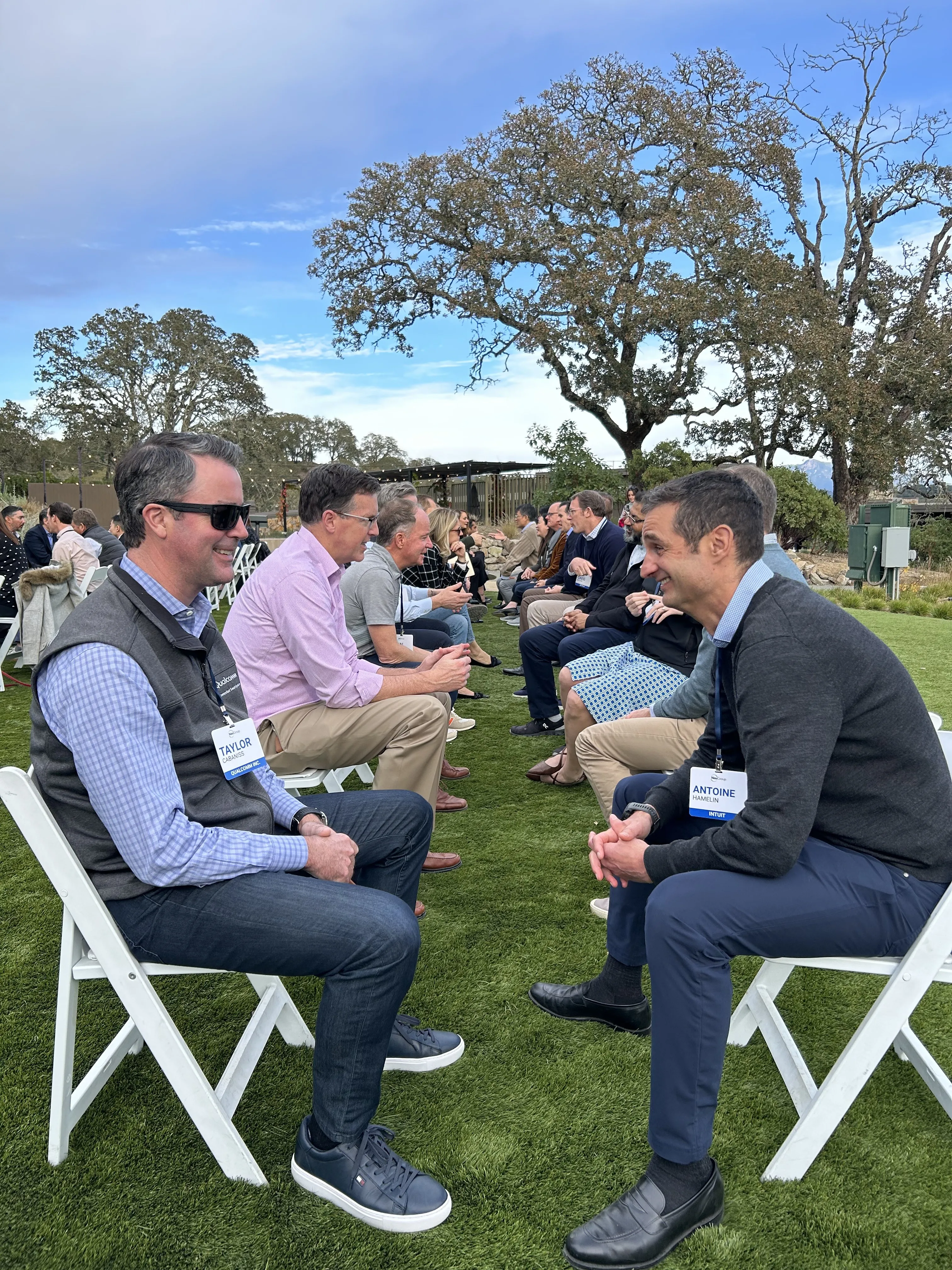 Two rows of people seated on white chairs facing each other outdoors on green grass, engaged in conversation with trees and a blue sky in the background.
