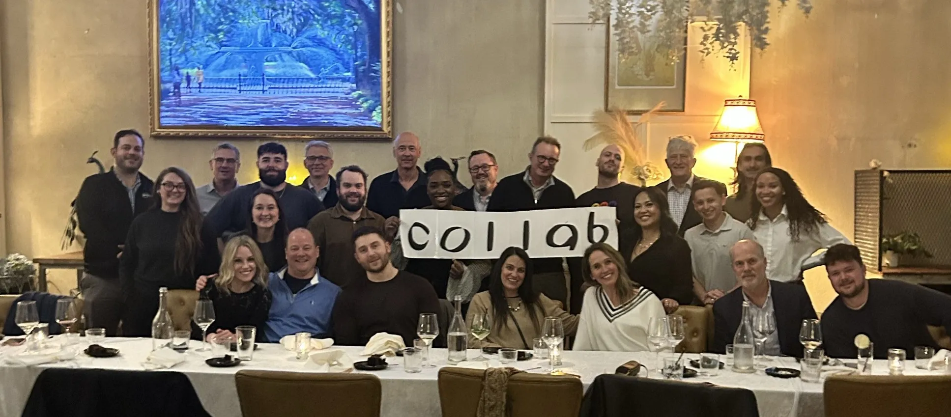 Group of 25 diverse people posing together indoors behind a long dining table, some holding signs spelling 'collab'.