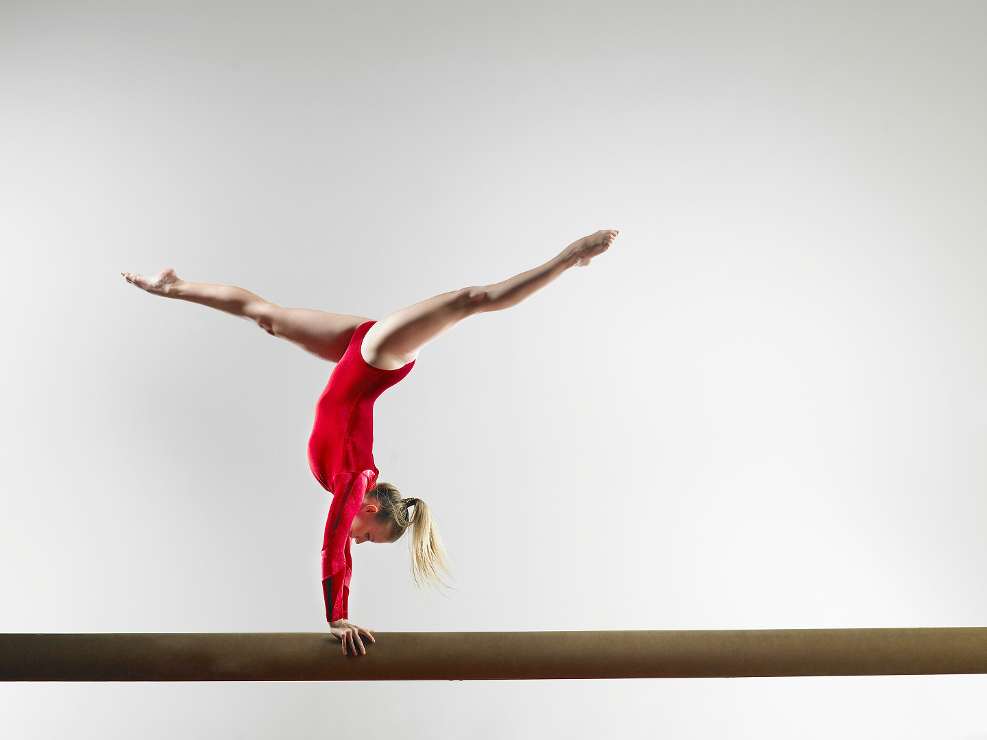 Female gymnast in a red leotard performing a handstand split on a balance beam.