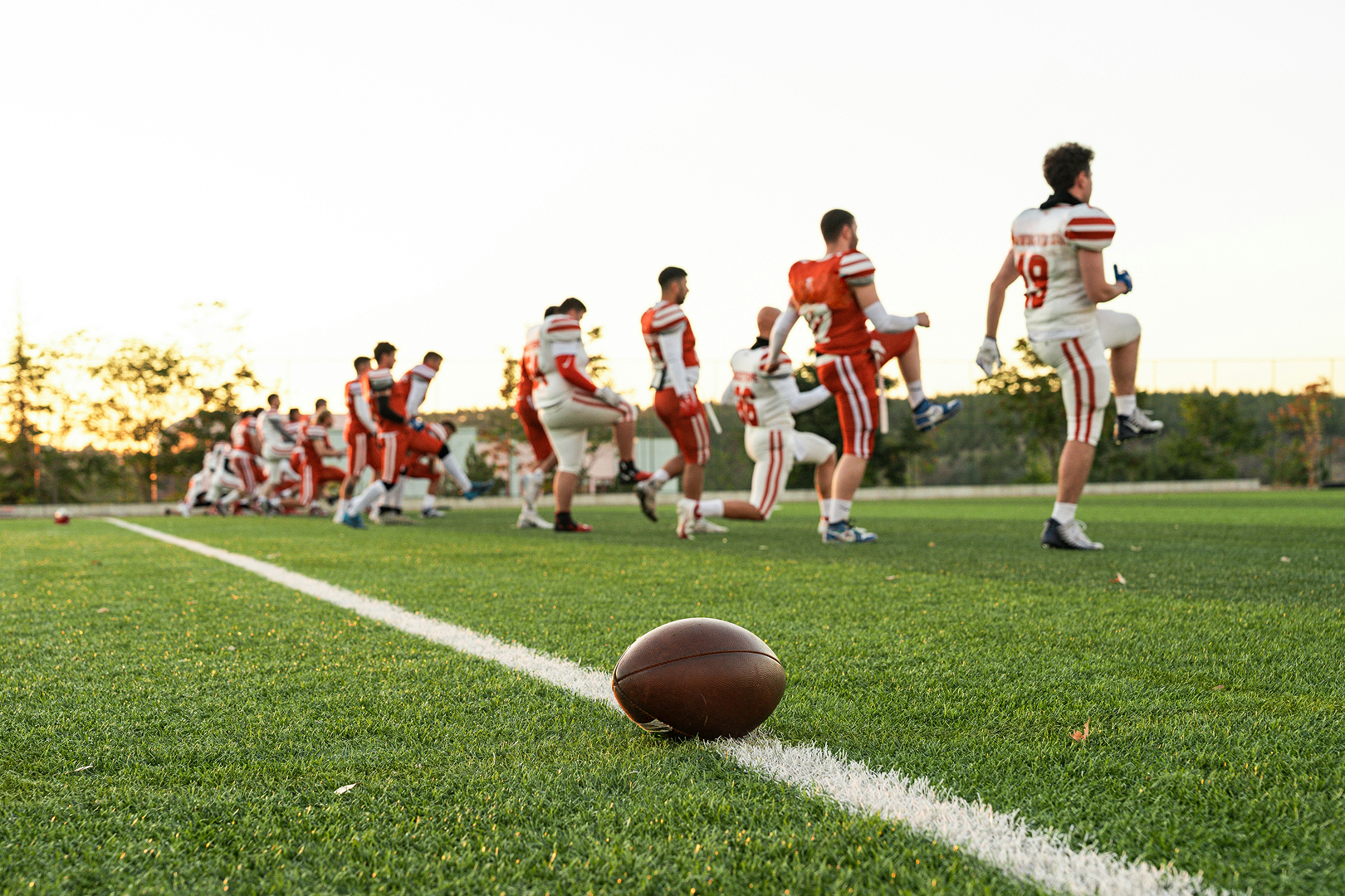 American football resting on green field with players in white and red uniforms stretching in the background.