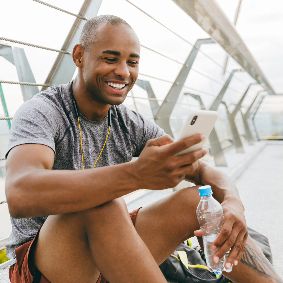 Smiling man in athletic wear sitting on the ground, holding a water bottle and looking at his smartphone.