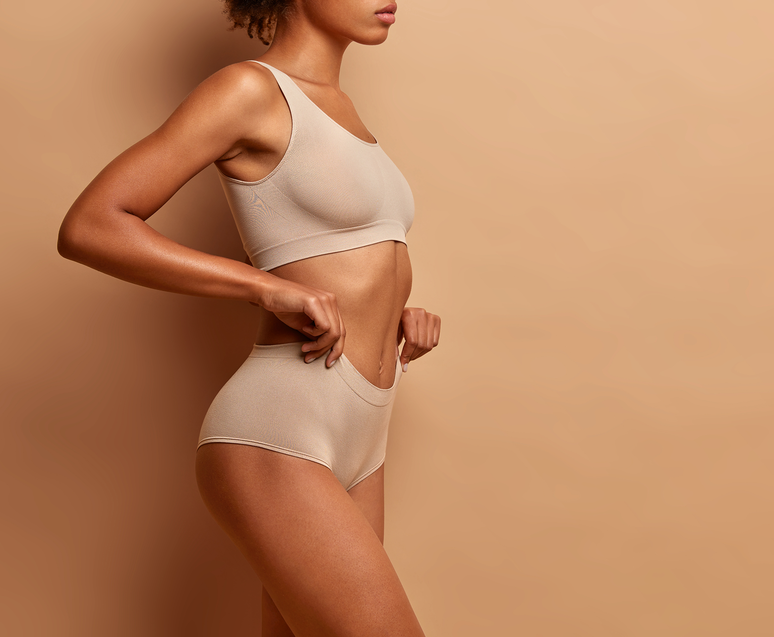 Woman in beige underwear pulling waistband of high-waisted briefs away from her waist against a tan background.