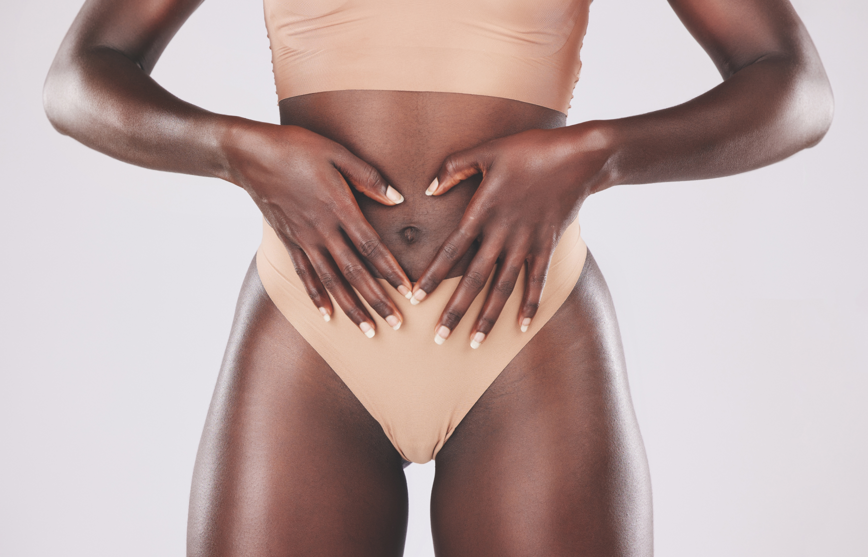 Close-up of a dark-skinned woman wearing beige underwear forming a heart shape with her hands on her stomach.