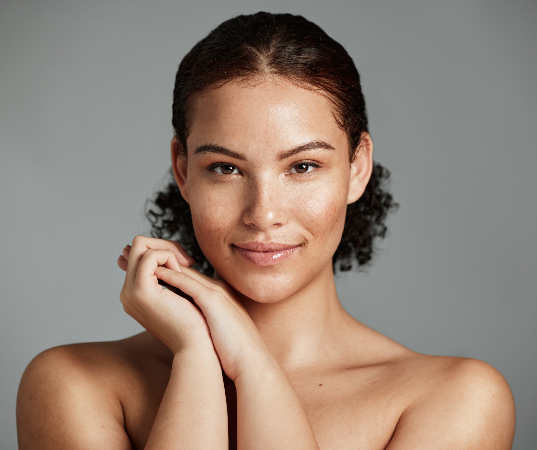 Close-up of a smiling woman with clear skin holding her hands near her face against a gray background.