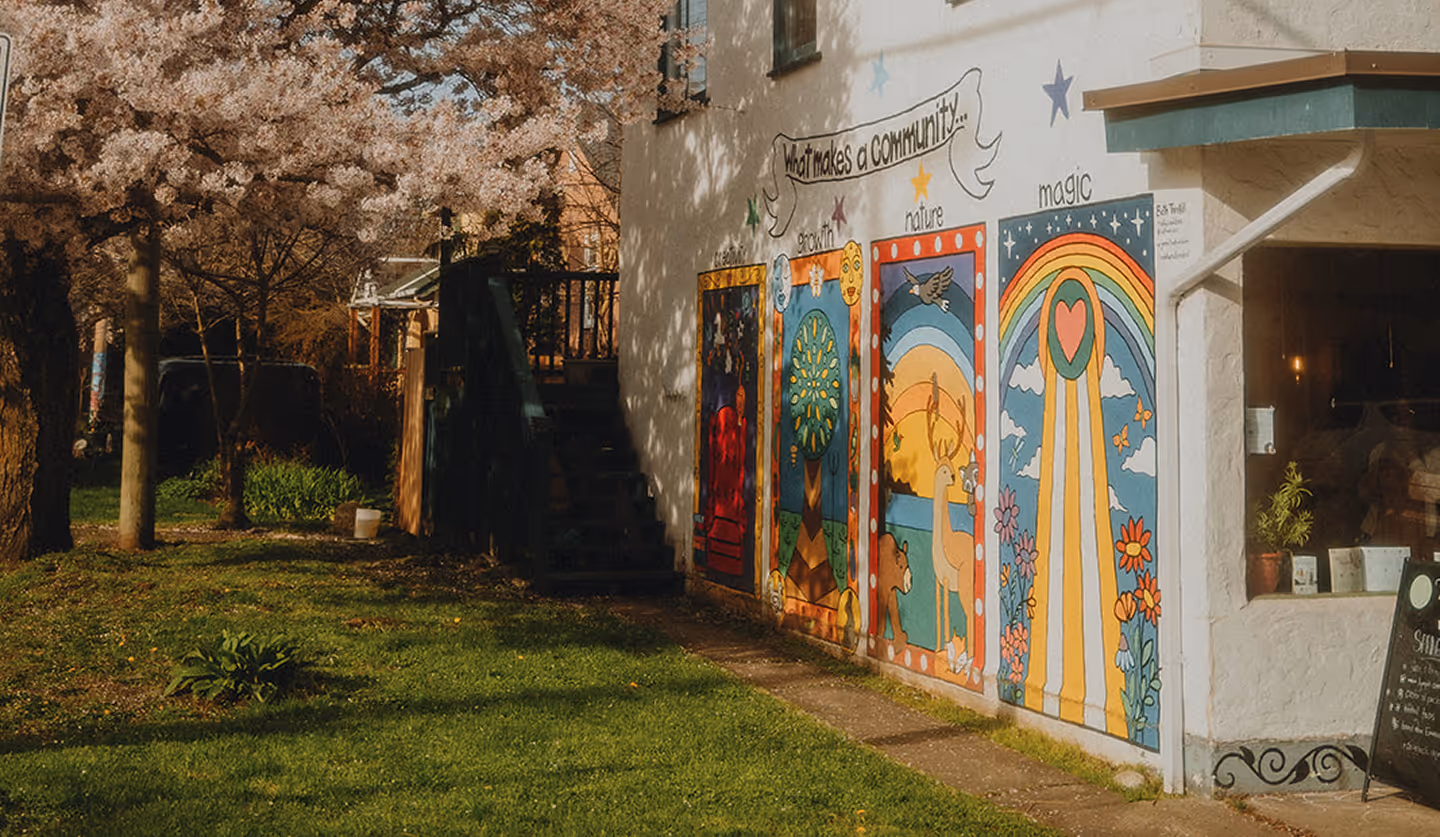 Colorful community-themed mural on a building wall beside a green lawn and blooming tree.