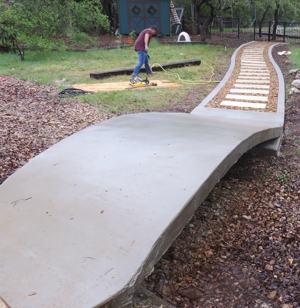 Curved concrete and stone pathway in a garden with a person working on wooden planks nearby.