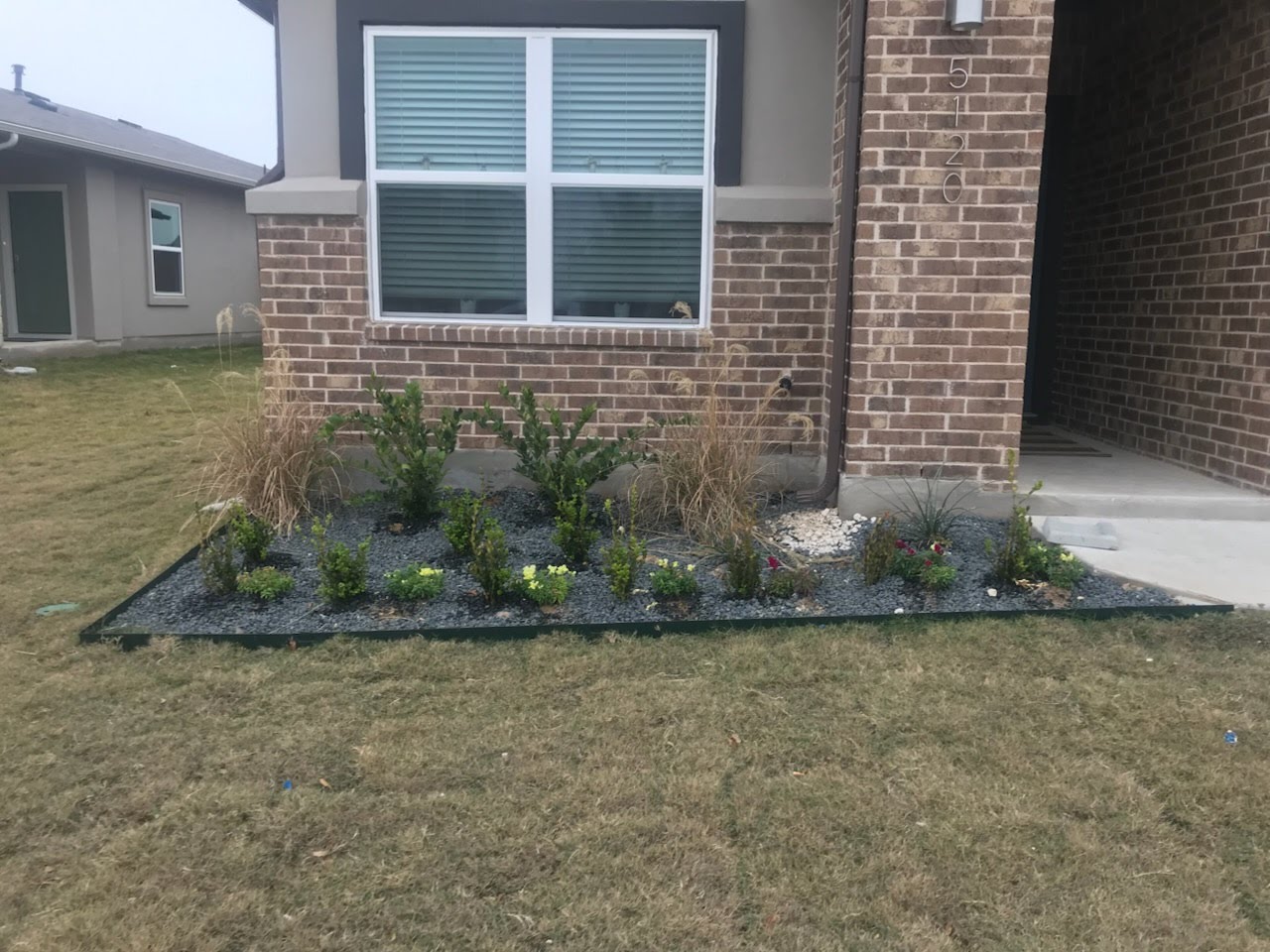 Person wearing gloves and a cap kneeling on grass while gardening next to lush green plants and a clear blue sky.