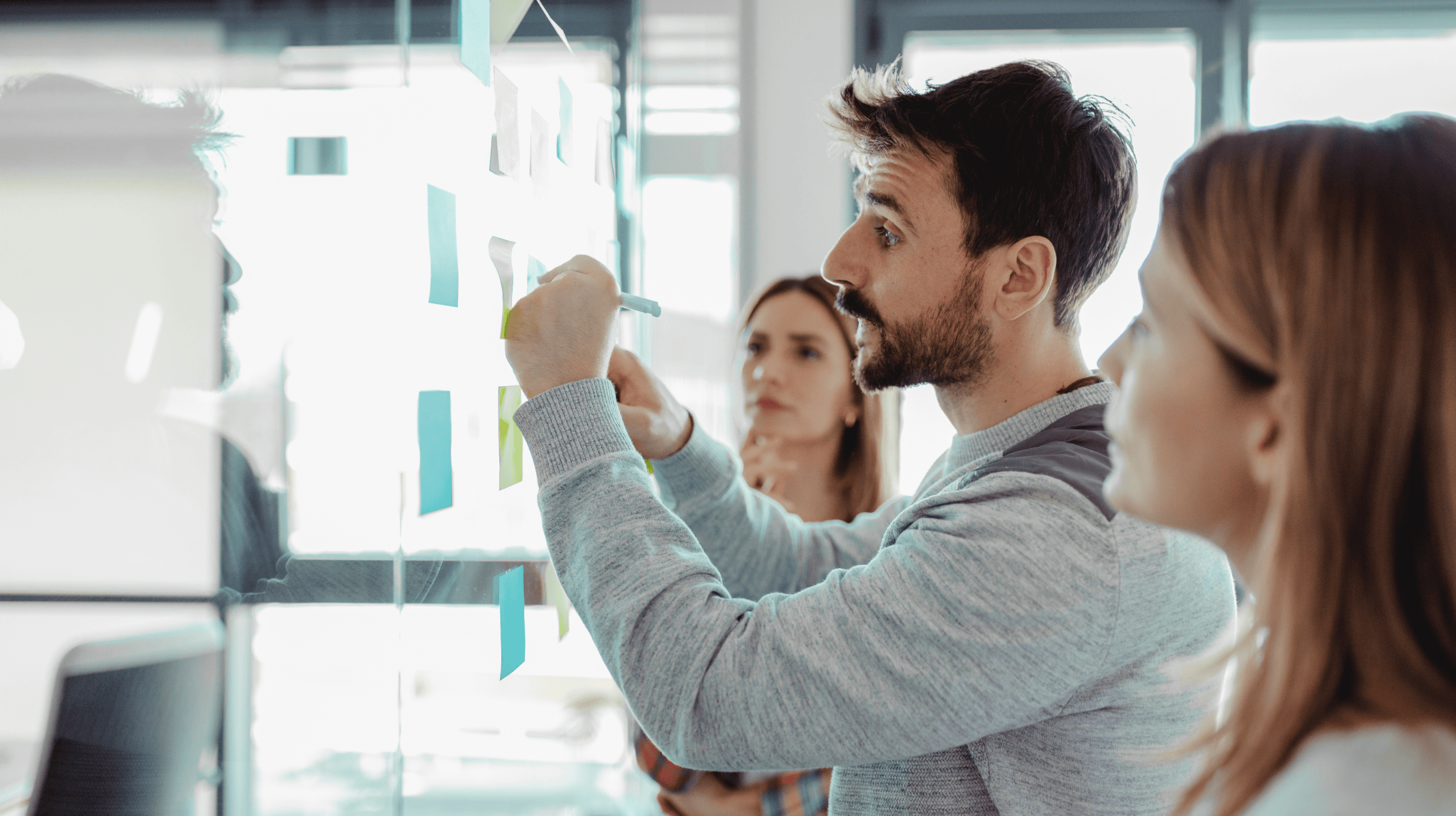 Three cybersecurity professionals, one man and two women, working through a security issue by posting sticky notes on a glass wall. Representative of the TPG Consulting website's homepage.