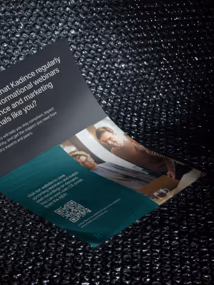 A folded brochure on a textured black surface showing text about Kadince webinars and a photo of two people smiling at a meeting.