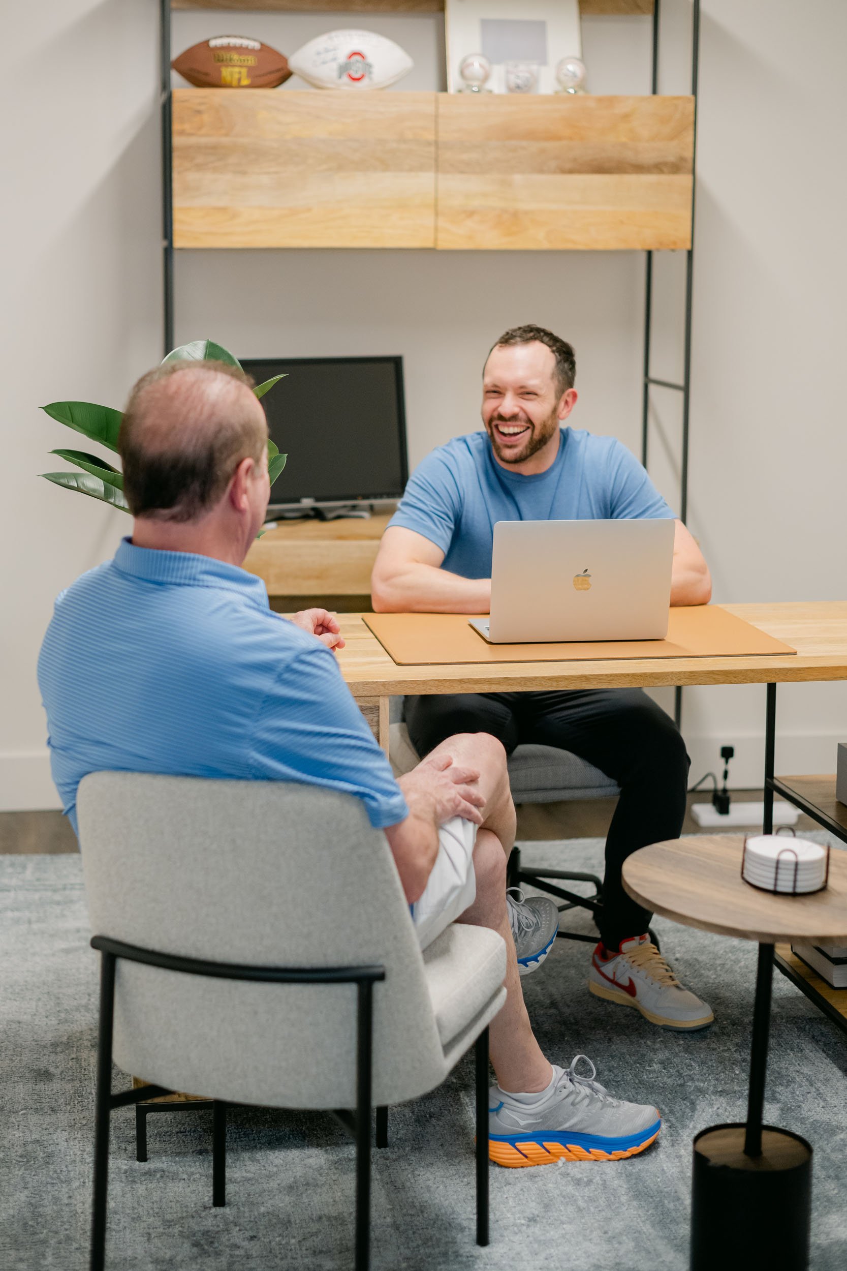 Two men having a friendly conversation in a modern office, one seated with a laptop smiling broadly.