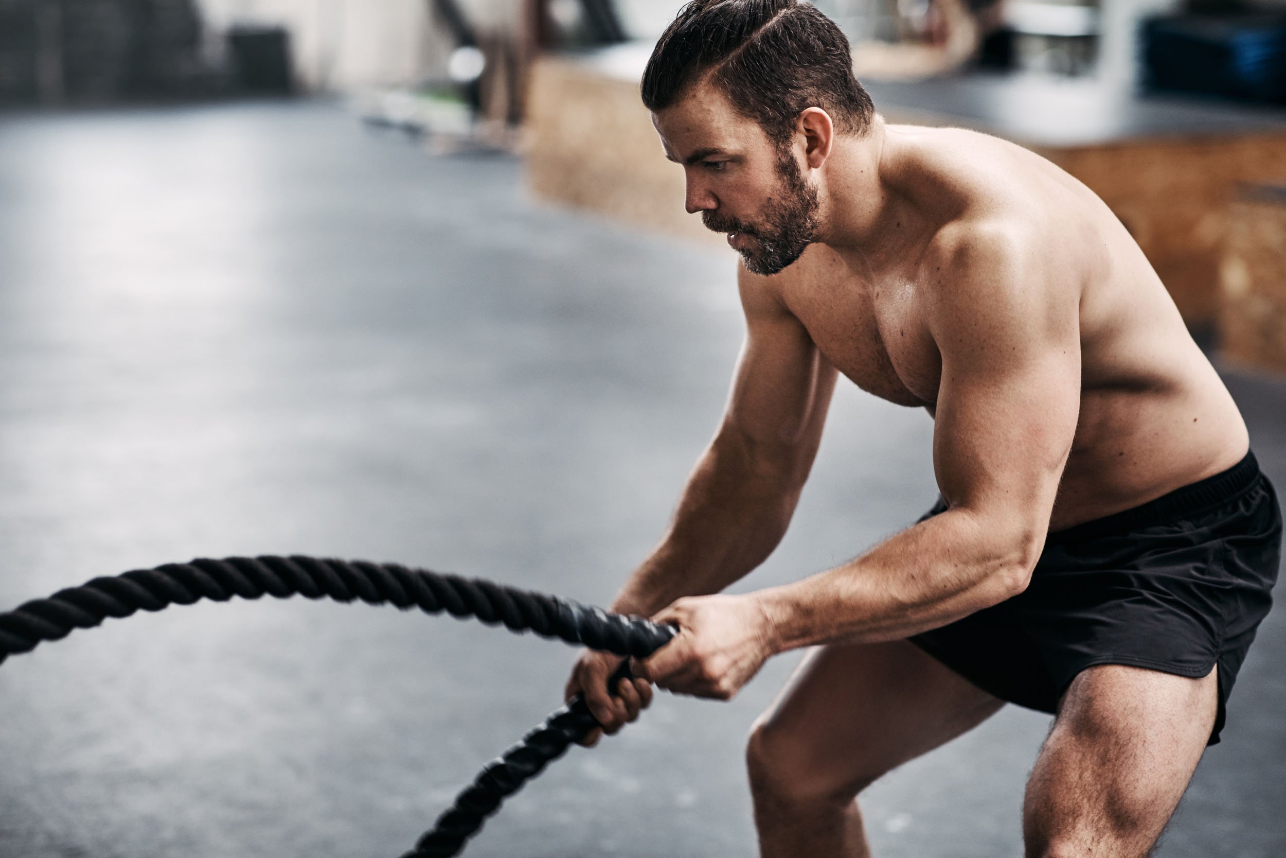 Shirtless man with beard intensely exercising with battle ropes in a gym.