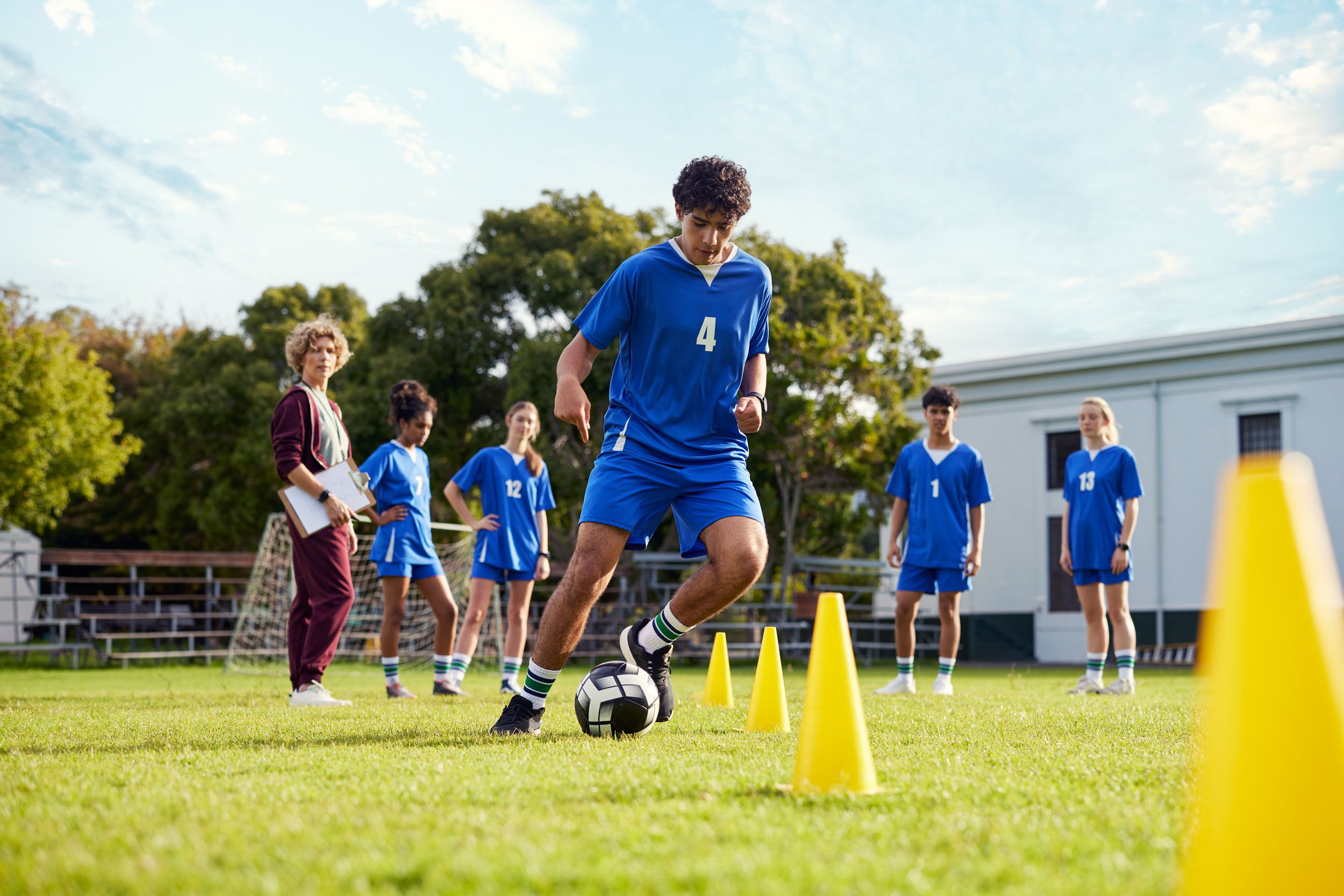 Young soccer player in blue jersey dribbling a ball through yellow cones during practice with teammates and coach observing.