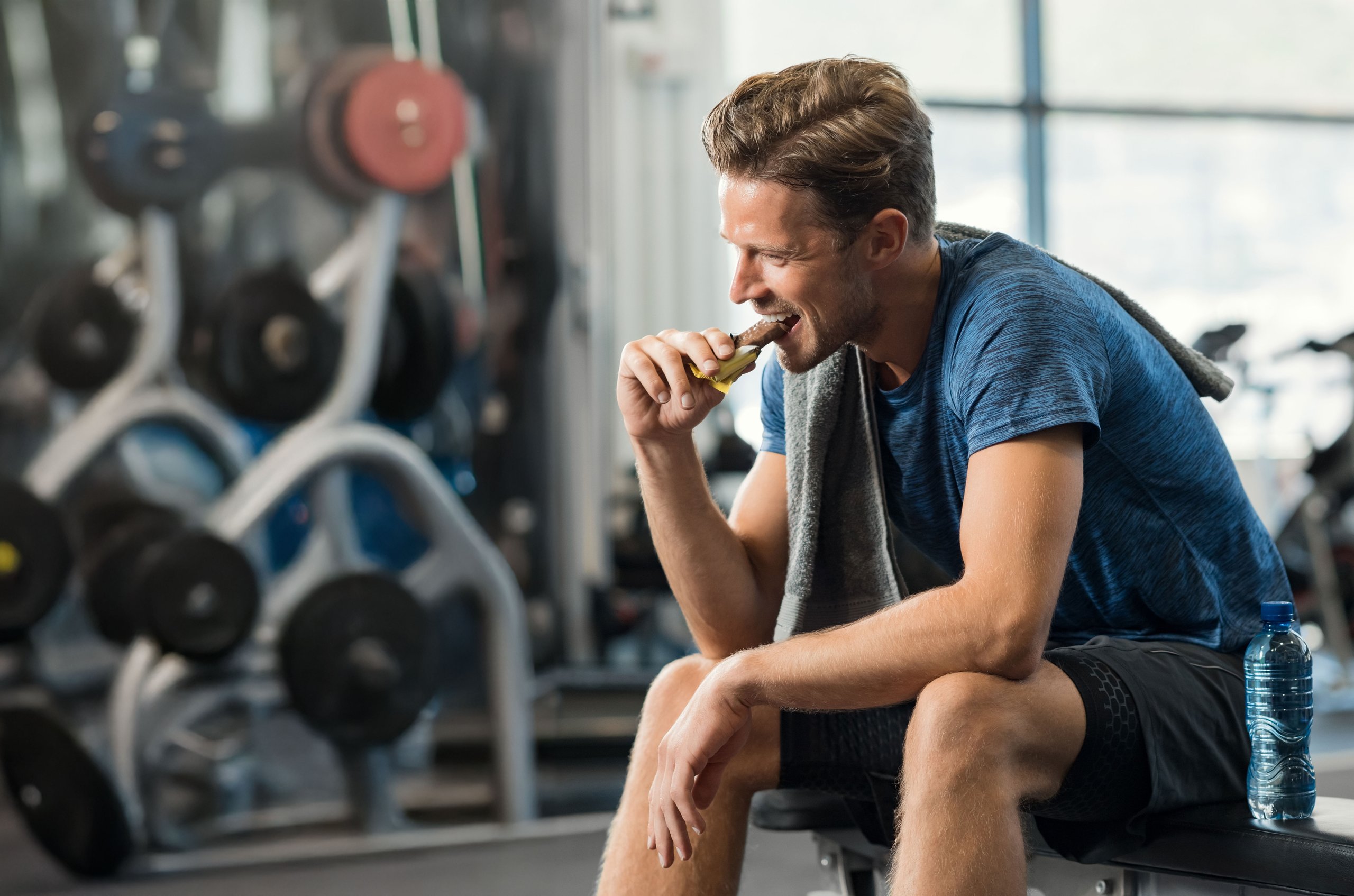 Man in workout clothes sitting on a gym bench eating a protein bar with a towel over his shoulder and a water bottle beside him.