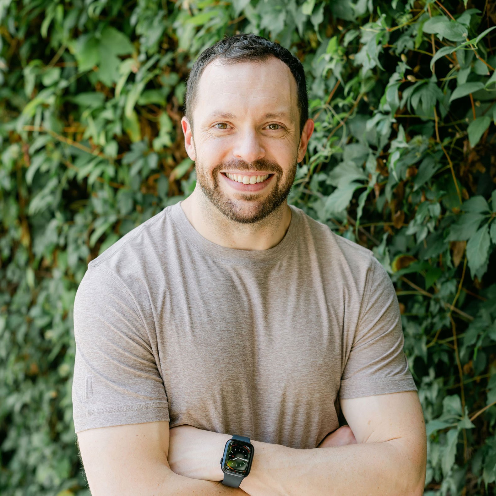 Smiling man with beard wearing a brown t-shirt and smartwatch, standing in front of green leafy background with arms crossed.