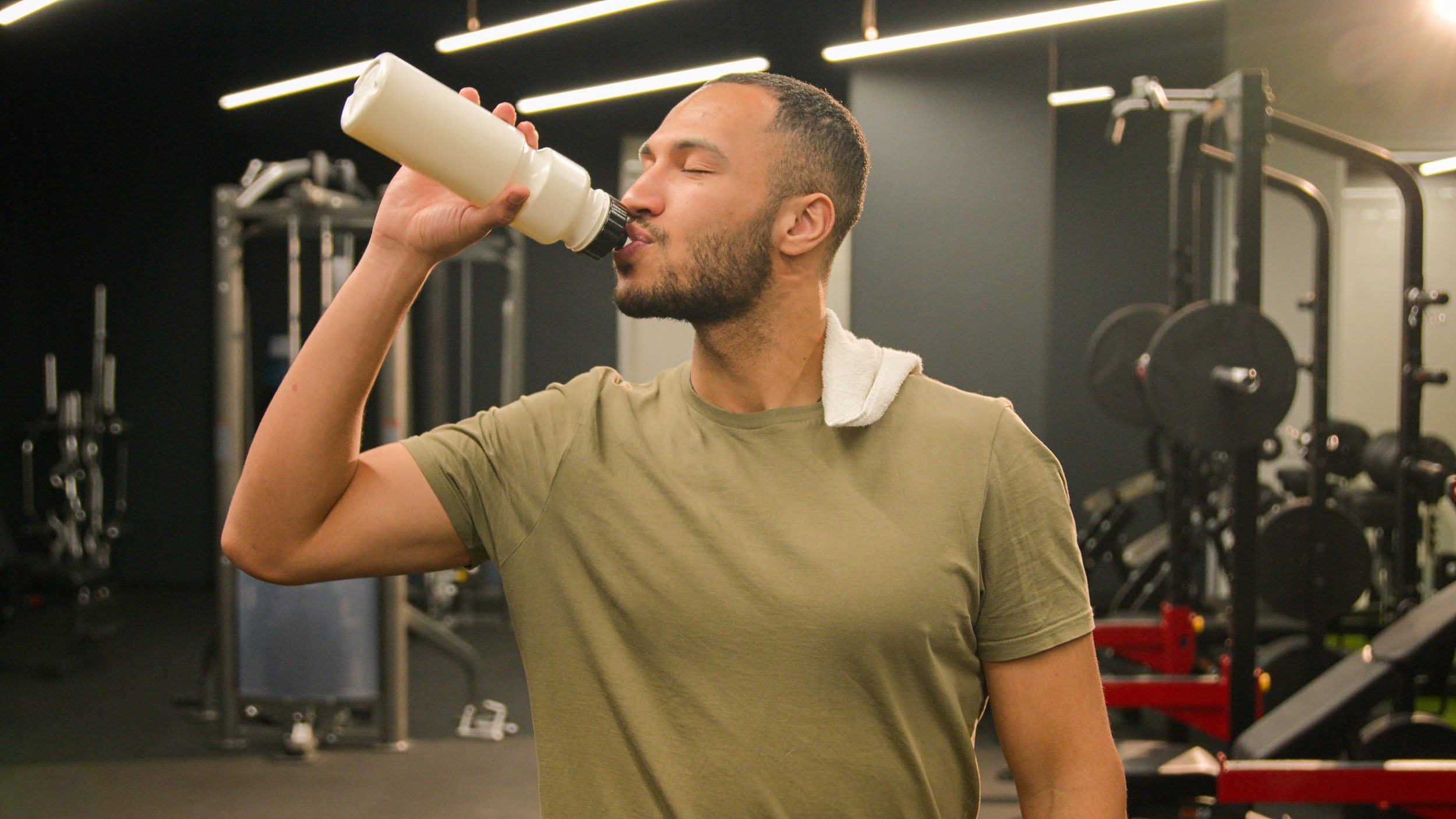 Man with a towel over his shoulder drinking water from a white bottle in a gym.