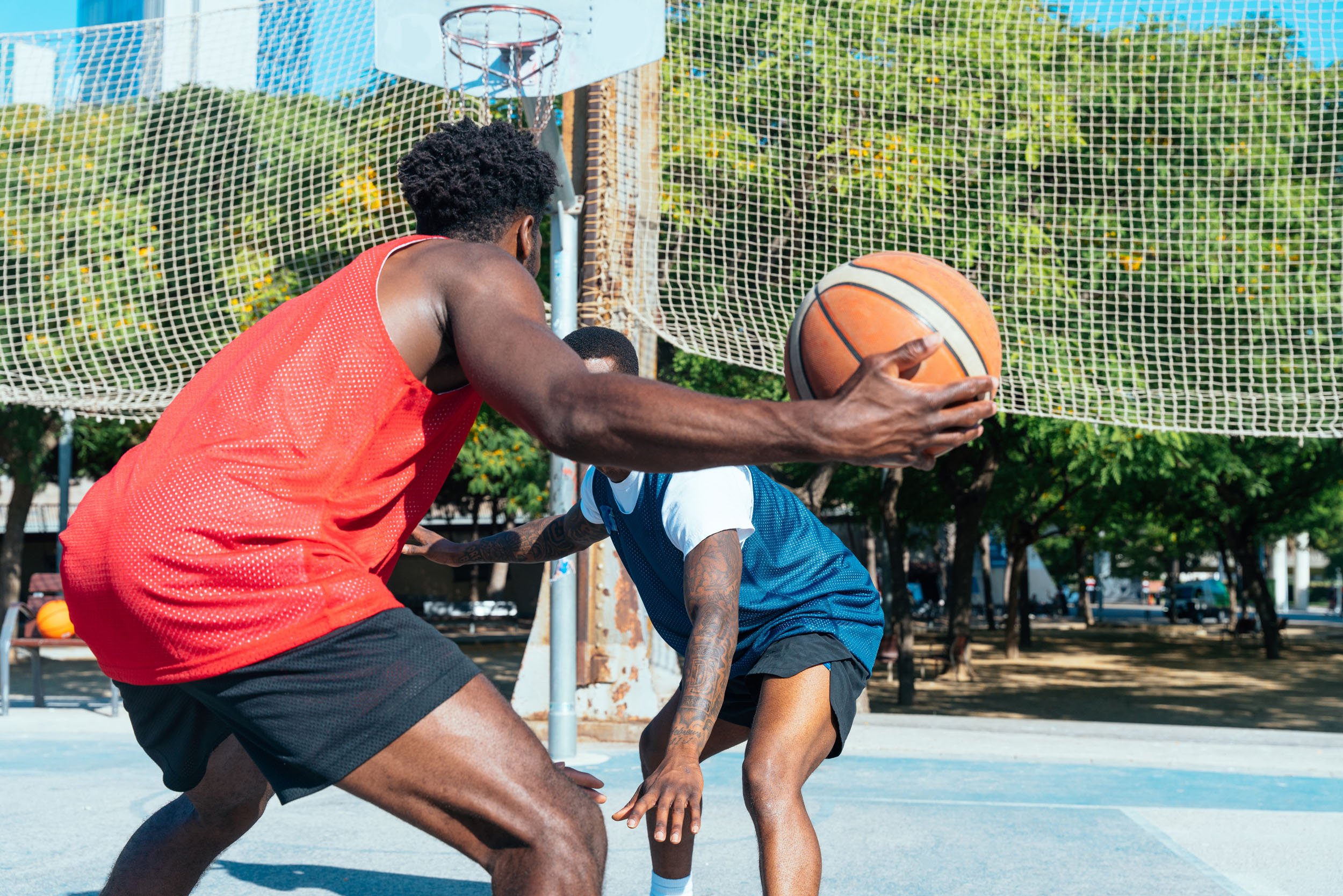 Two men playing basketball on an outdoor court, one wearing a red jersey holding the ball, the other in a blue jersey defending.