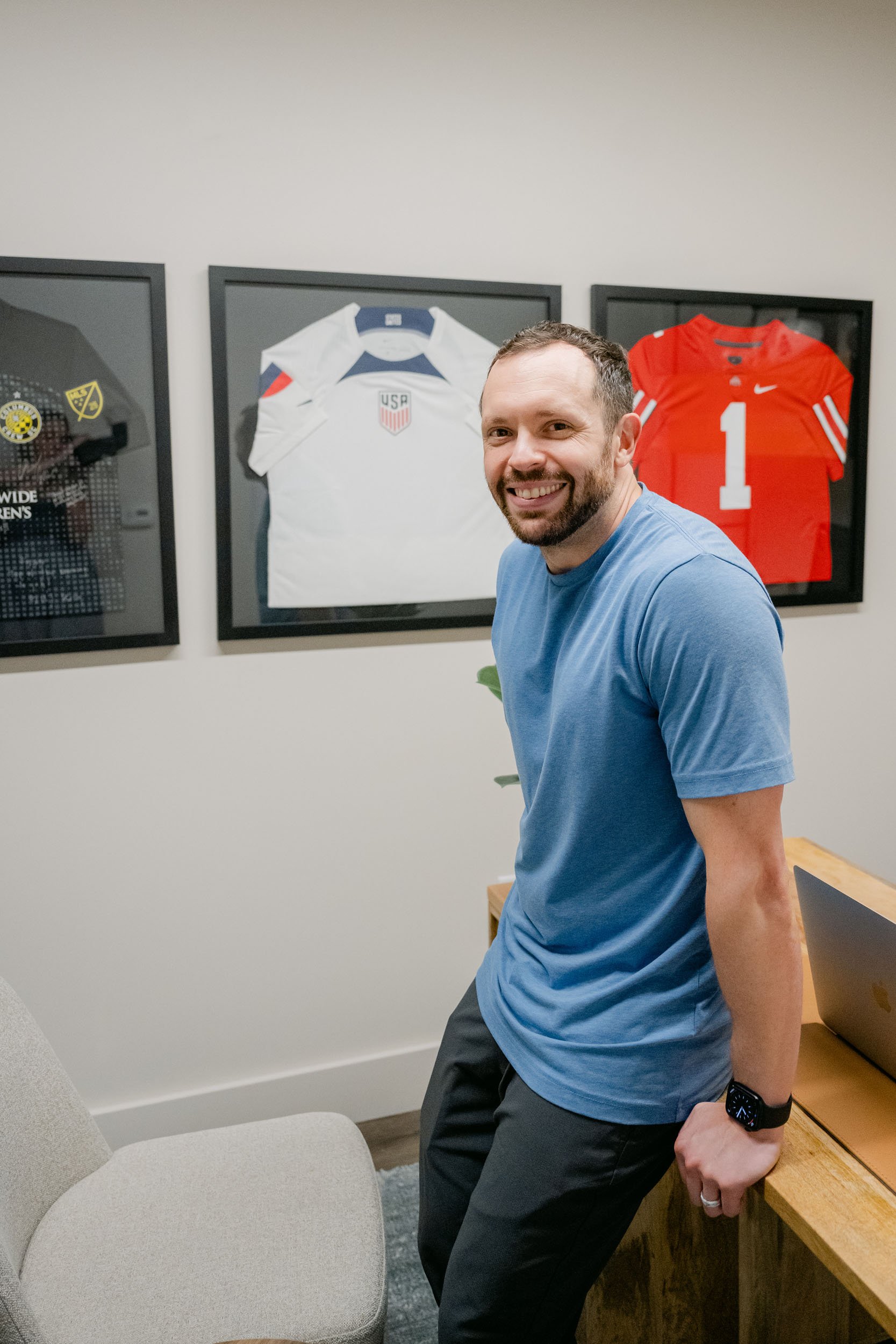 Smiling man in a blue t-shirt leaning against a wooden desk in a room with three framed sports jerseys on the wall.