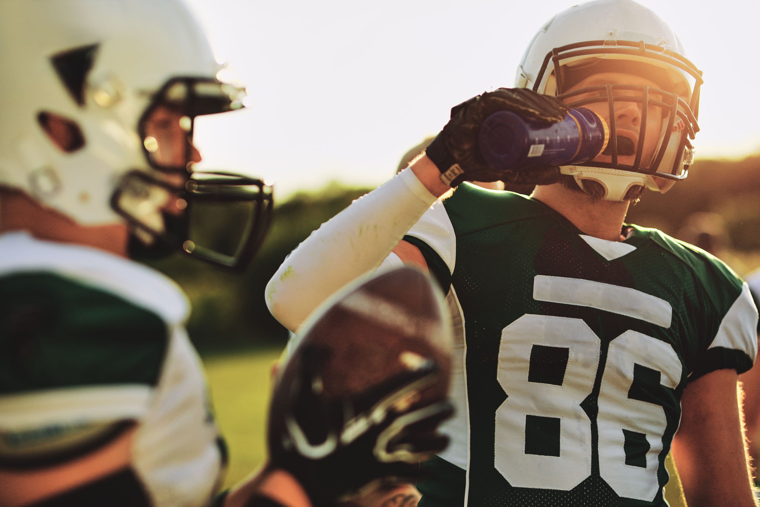 American football player wearing jersey number 86 drinks from a water bottle while teammates stand nearby on a sunlit field.