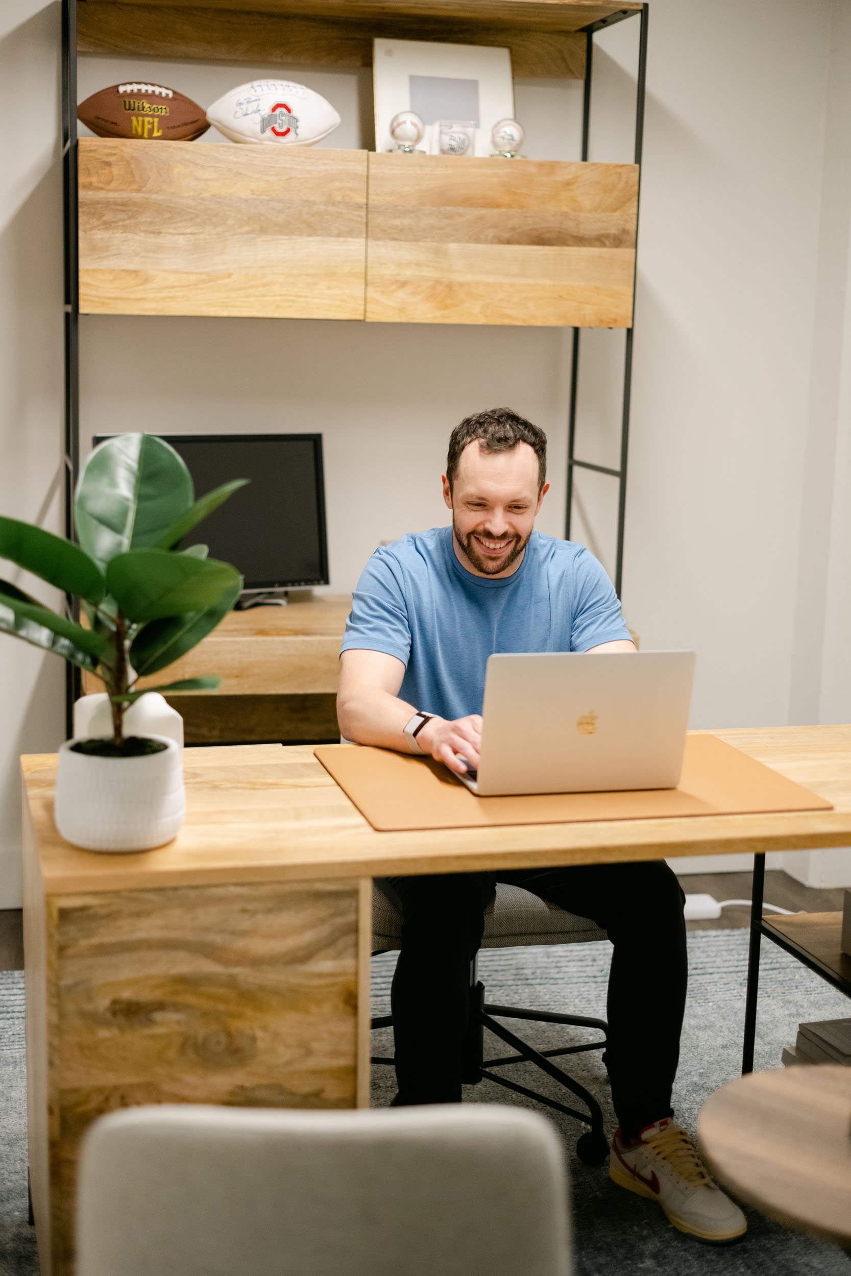 Man in blue shirt smiling while working on a laptop at a wooden desk in a modern office with sports memorabilia on shelves behind.