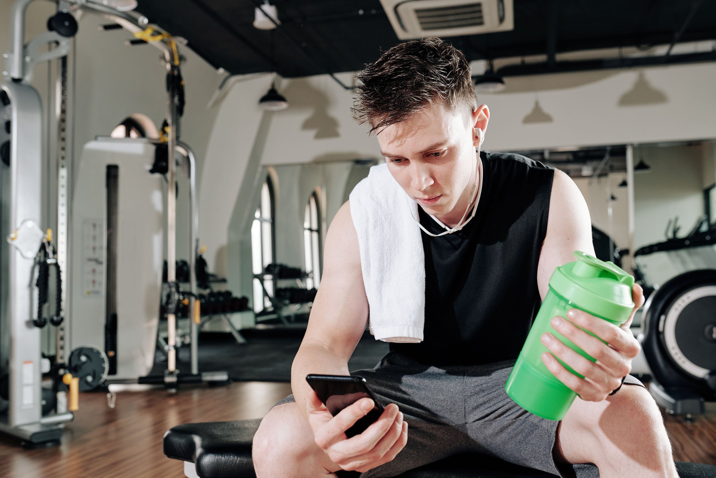 Young man in gym gear sitting on bench holding a green shaker bottle and looking at his smartphone with a white towel over his shoulder.
