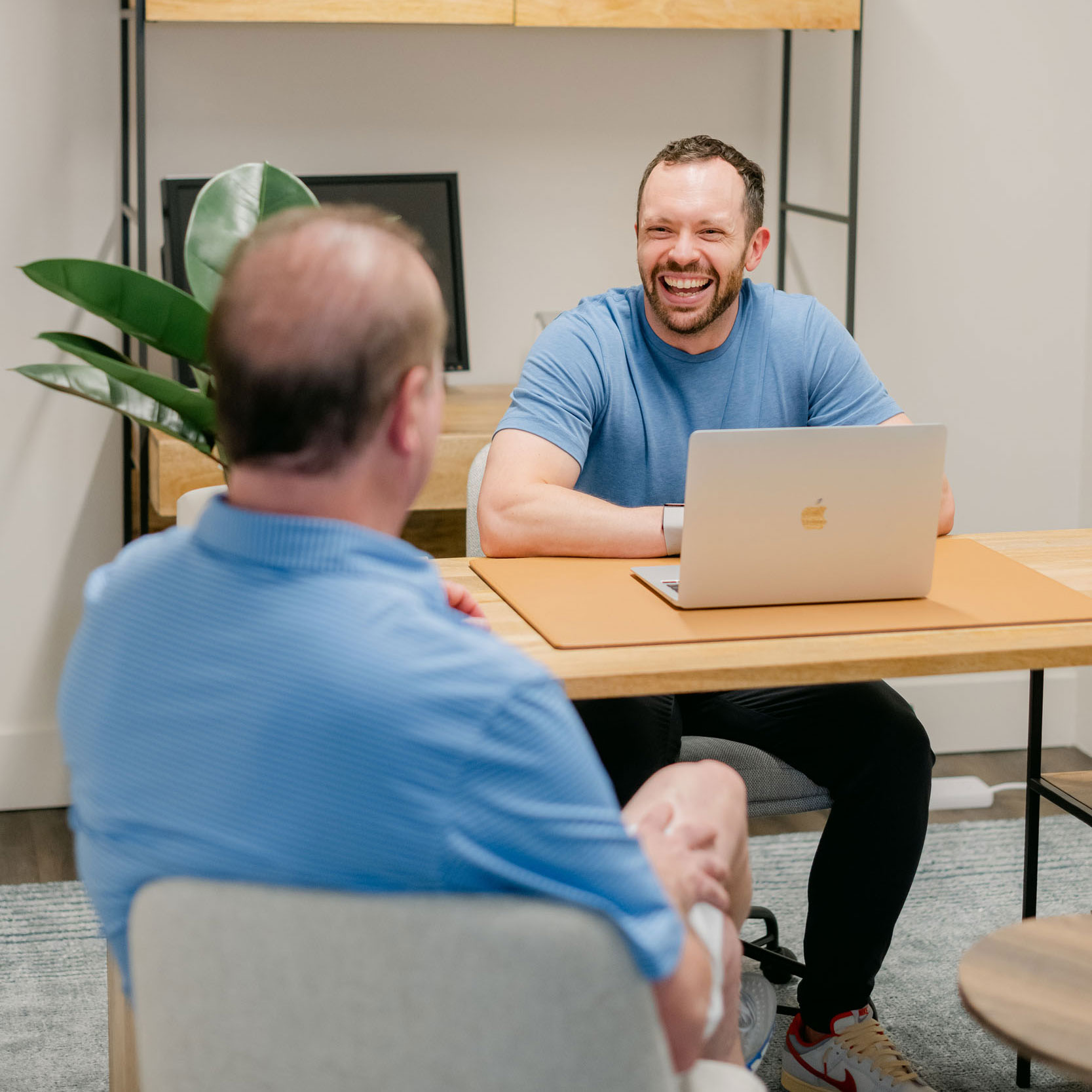 Two men sitting across from each other at a wooden table, one smiling with a laptop in front of him.