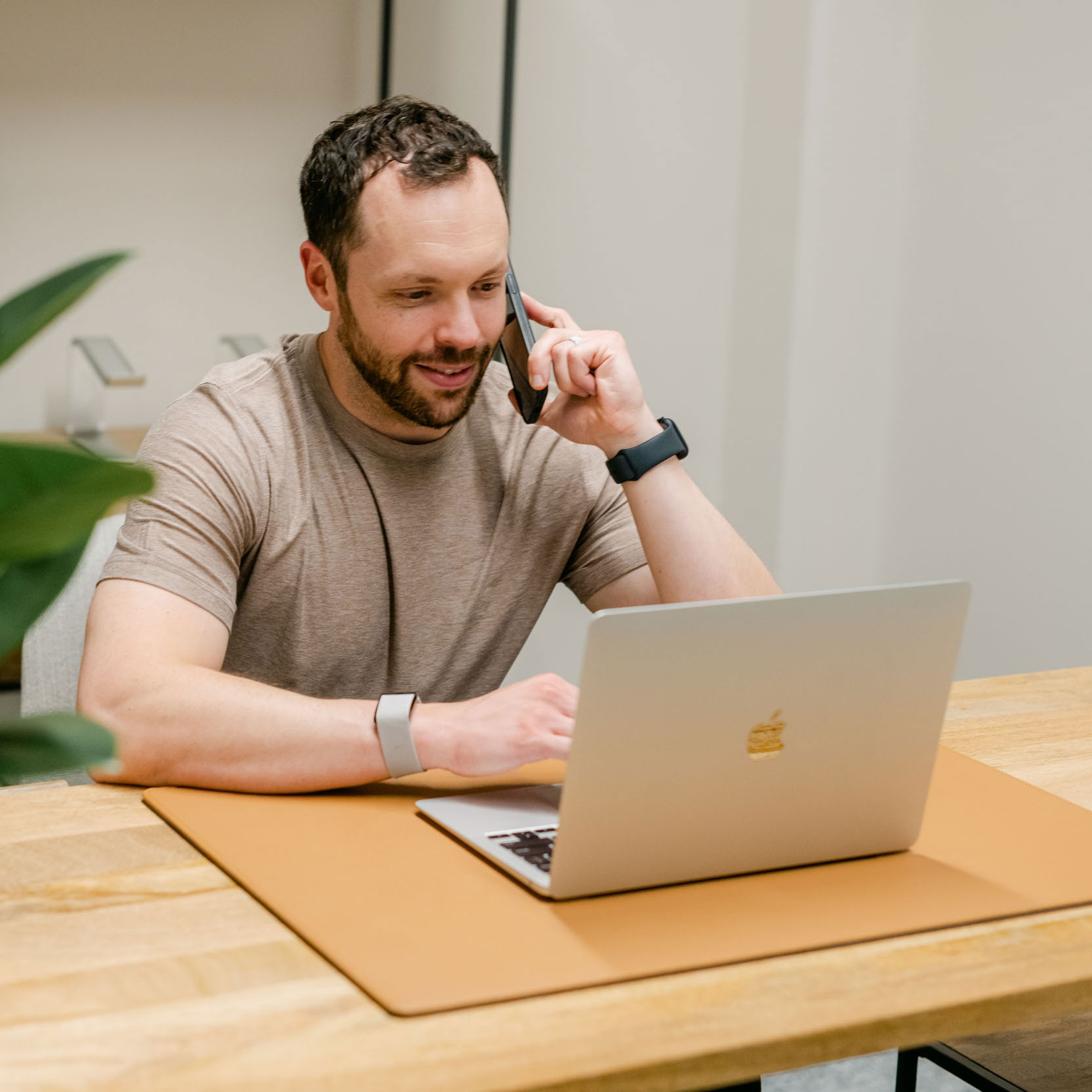 Man in a beige t-shirt talking on a smartphone while working on a laptop at a wooden table.