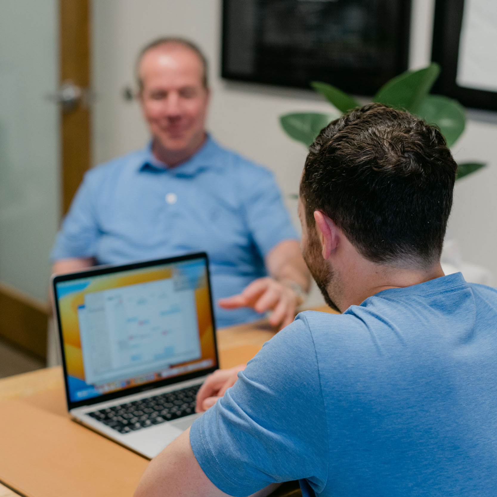 Two men in blue shirts sitting at a table, one using a laptop with a calendar application open.