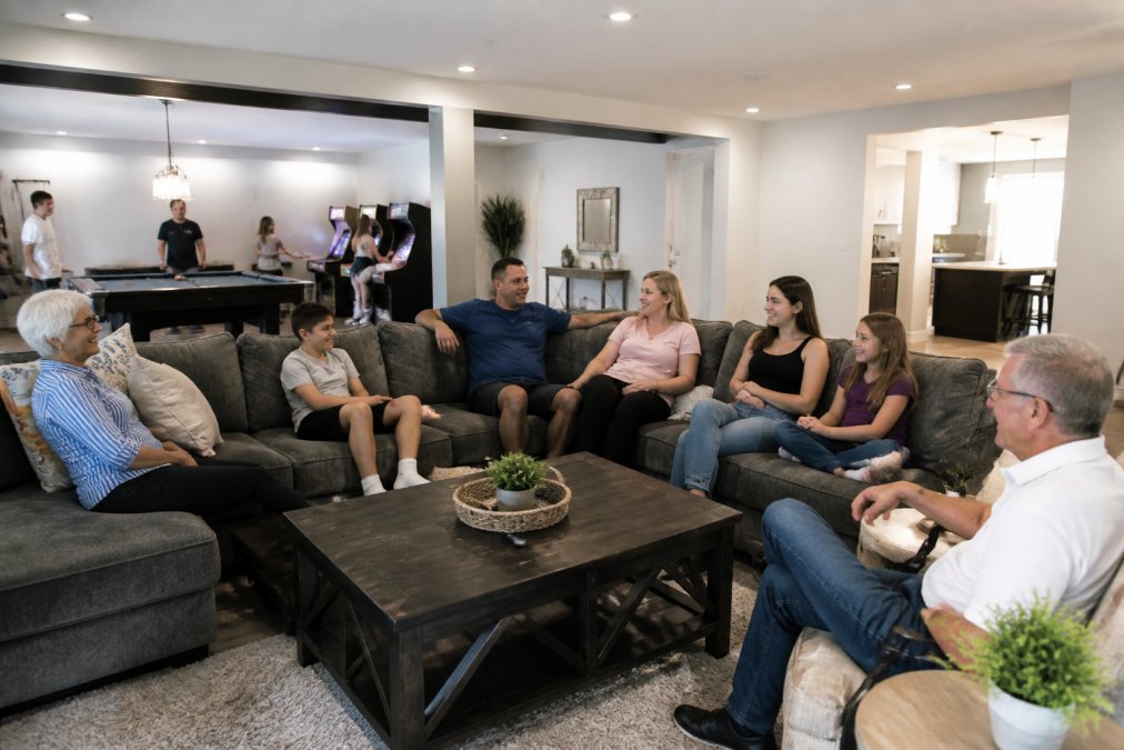 A spacious, well-lit living room in a modern vacation home with comfortable sectional seating, a pool table and arcade games visible in the adjacent game room, and a diverse multi-generational family relaxing together