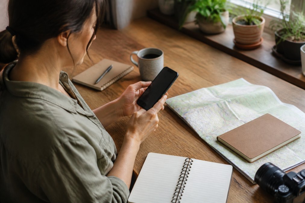 Person using smartphone to complete vacation rental booking while sitting at desk with travel planning materials and coffee nearby, warm natural lighting