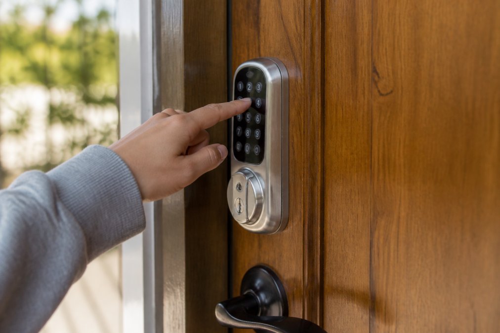 Close-up of a guest's hand entering a numeric code on a silver digital keypad mounted on a wooden front door, with soft afternoon sunlight illuminating the entry