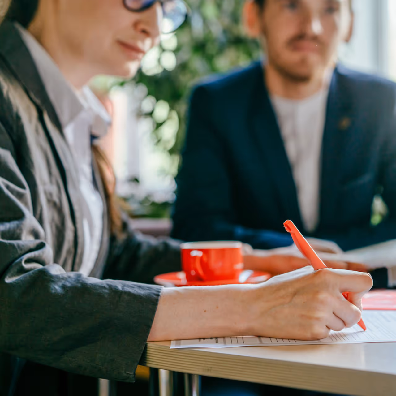 Person writing on a document with a red pen at a table, with a red cup and another person blurred in the background.