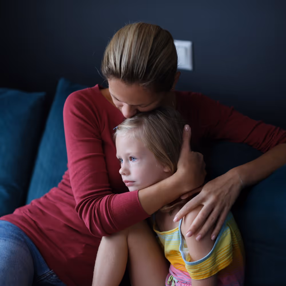 Woman wearing a red sweater comforting and hugging a sad young girl with blonde hair sitting on a blue couch.