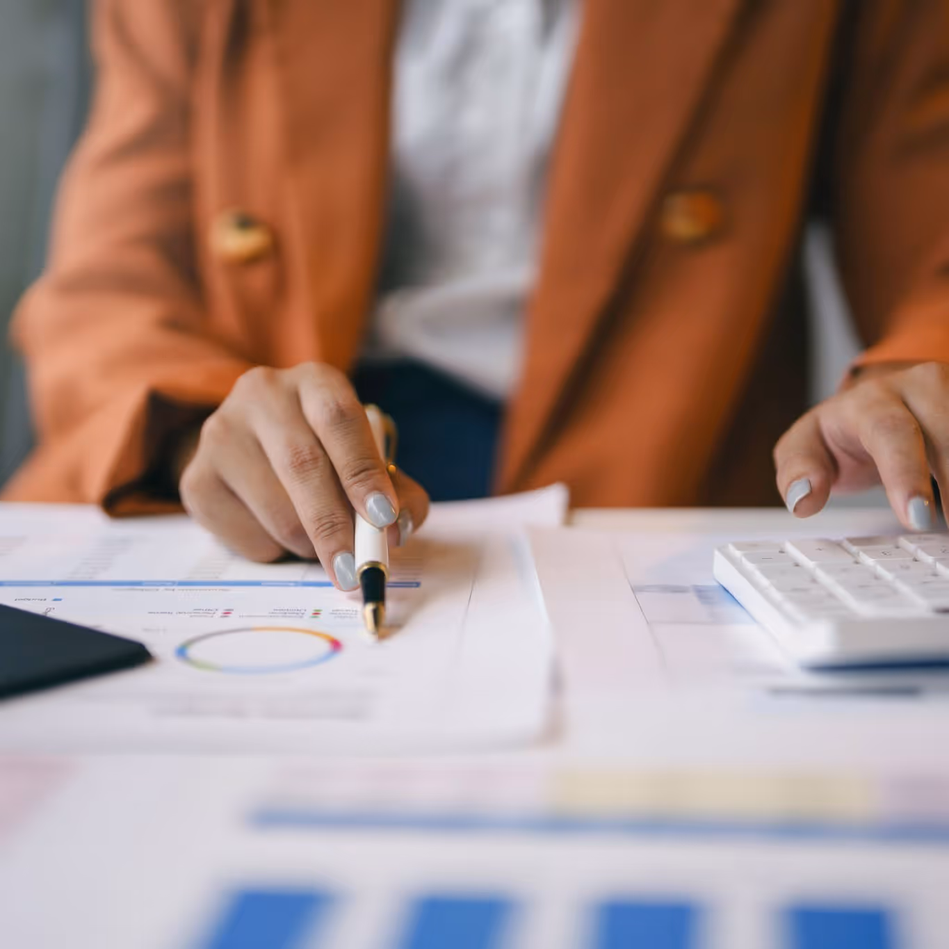 Person in brown jacket using a calculator and holding a pen over financial charts on a desk.