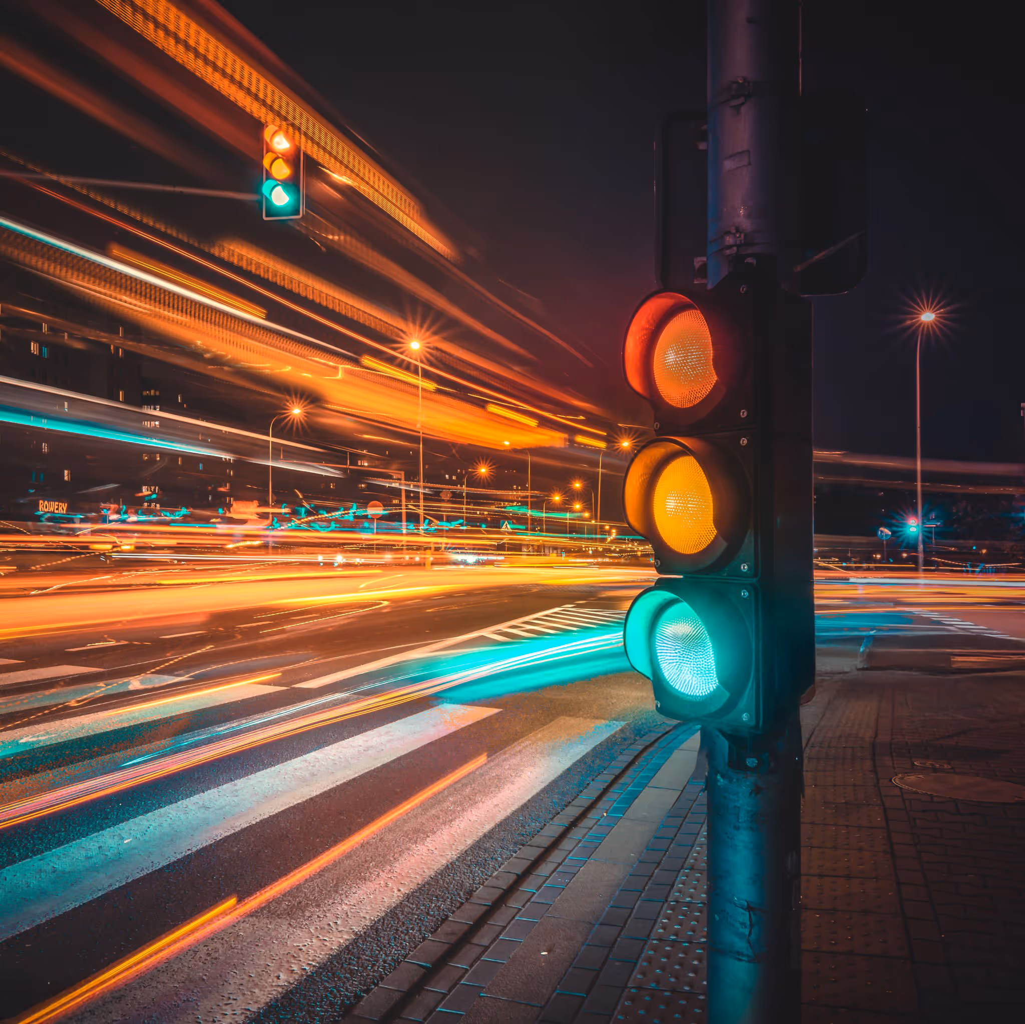 Traffic light at night showing red, yellow, and green lights with colorful light trails from moving vehicles on the road.