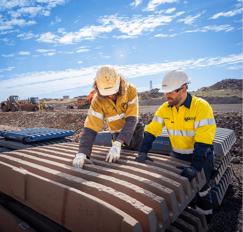 Smiling middle-aged man in a yellow high-visibility work shirt and hard hat holding a clipboard outdoors at a mining or construction site.