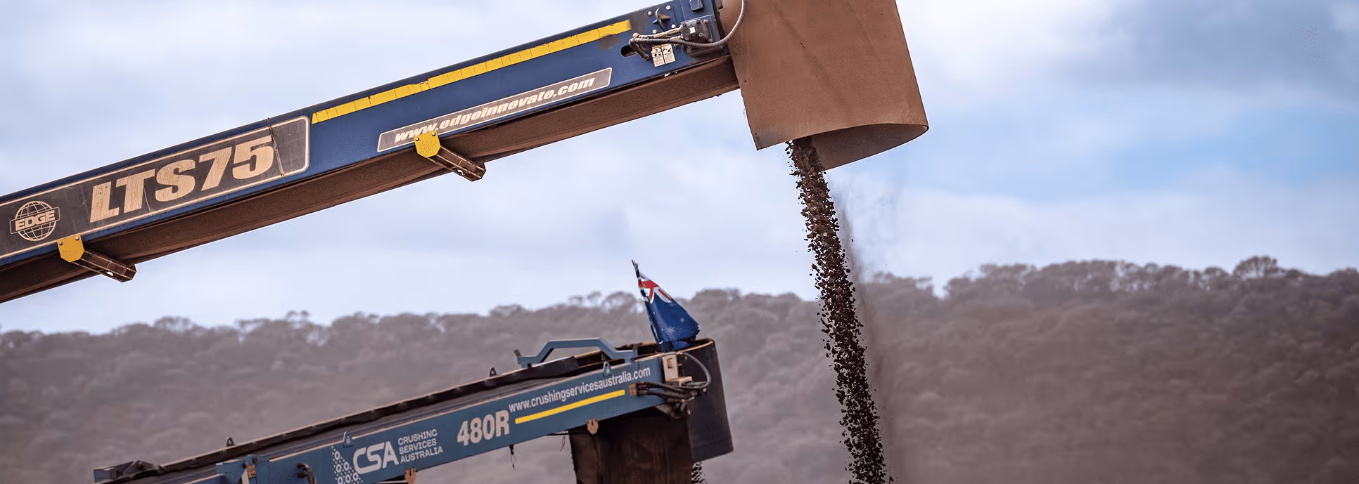 Conveyor belt machine unloading crushed rock with a landscape of trees and cloudy sky in the background.
