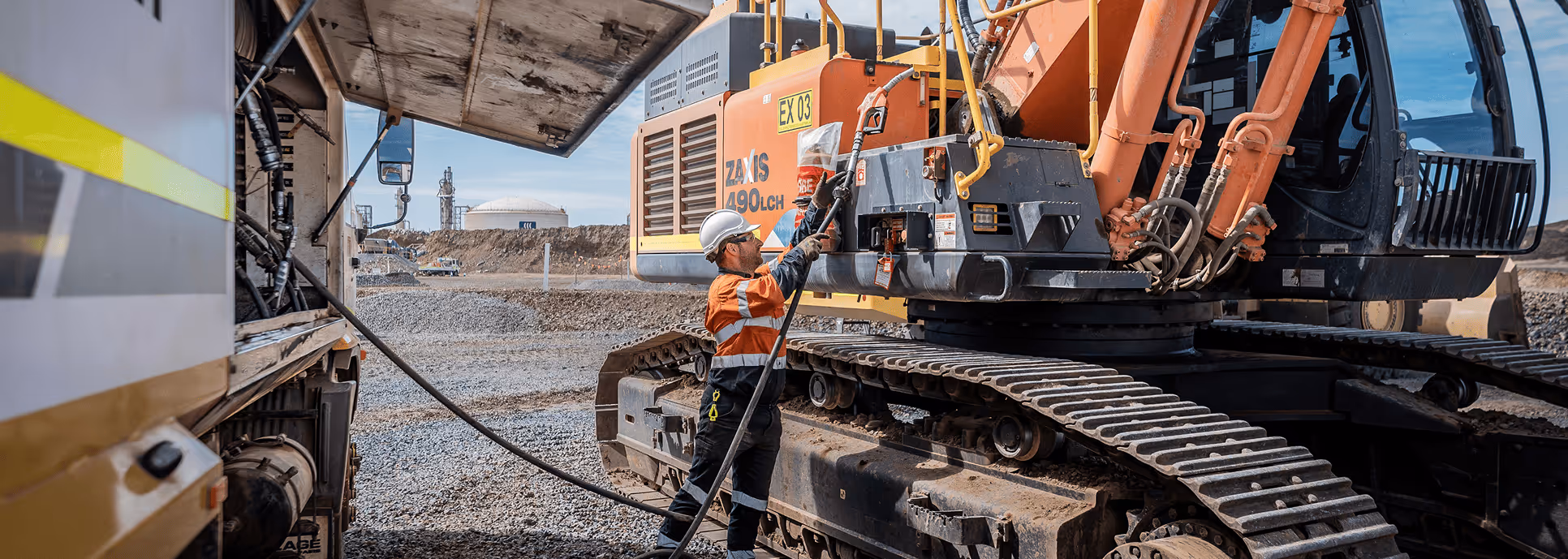 Worker in safety helmet and orange reflective jacket refueling a large excavator on a gravel construction site.