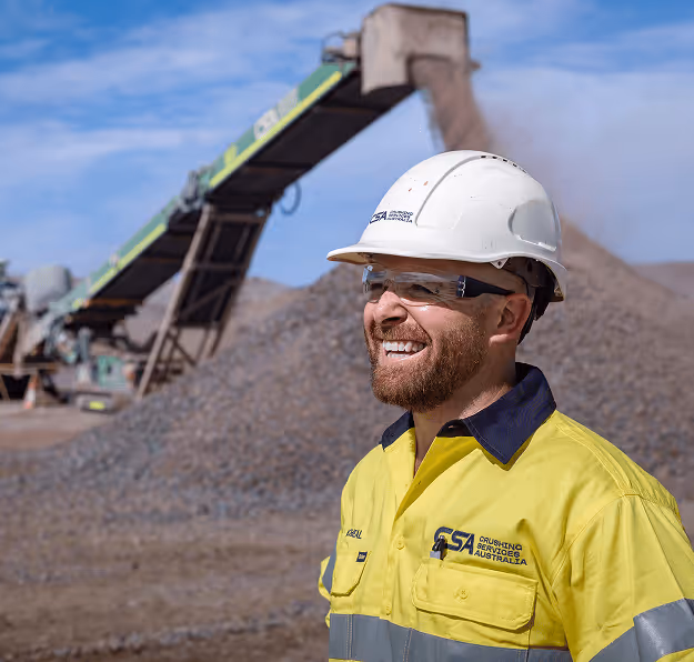 Smiling construction worker in yellow high-visibility jacket, white hard hat, and safety glasses at a gravel processing site.