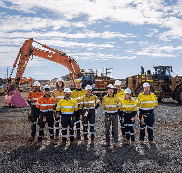 Group of nine miners and construction workers wearing high-visibility safety gear and helmets standing on gravel in front of large construction equipment under a partly cloudy sky.