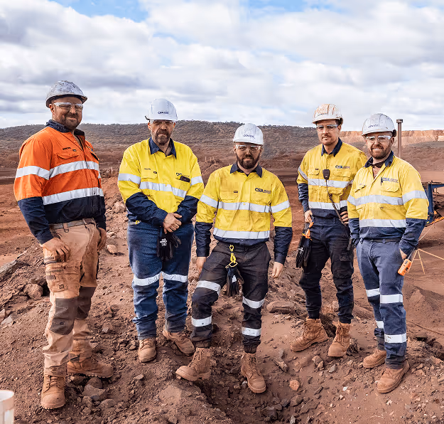 Five men wearing high-visibility work clothing and safety helmets standing on rocky ground at a mining or construction site.