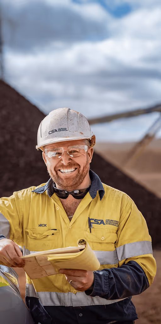 Smiling construction worker wearing a white hard hat and yellow safety jacket holding a clipboard outdoors.