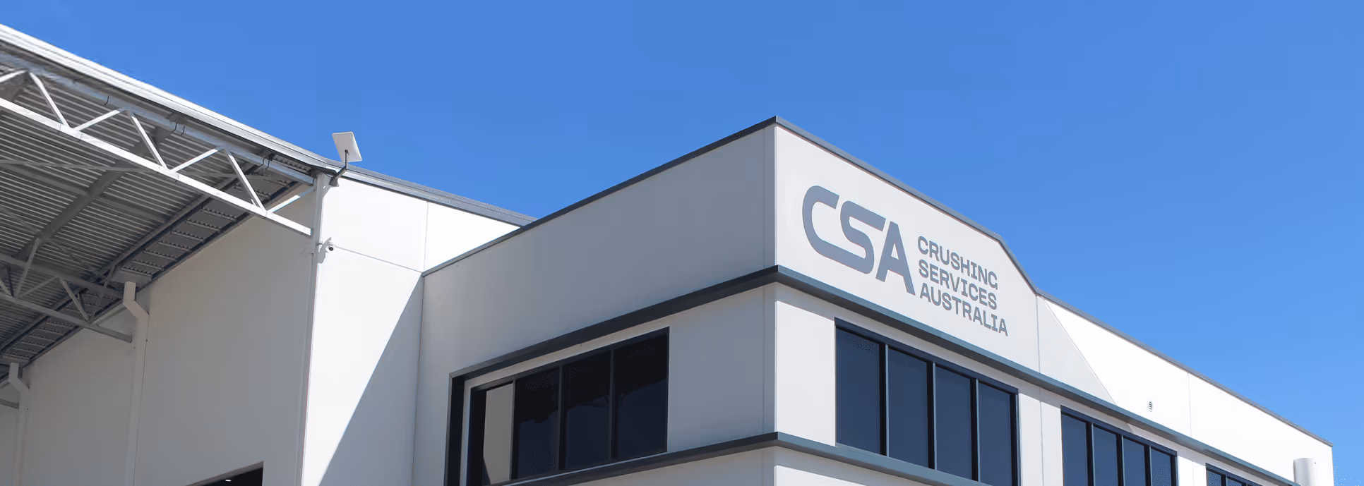 Modern white industrial building with large windows and CSA Crushing Services Australia logo on the upper corner under a clear blue sky.