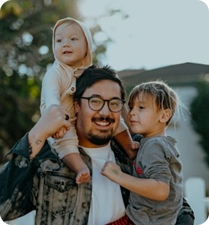 Smiling man with glasses holding a toddler on his shoulders and hugging a young child two outdoors.