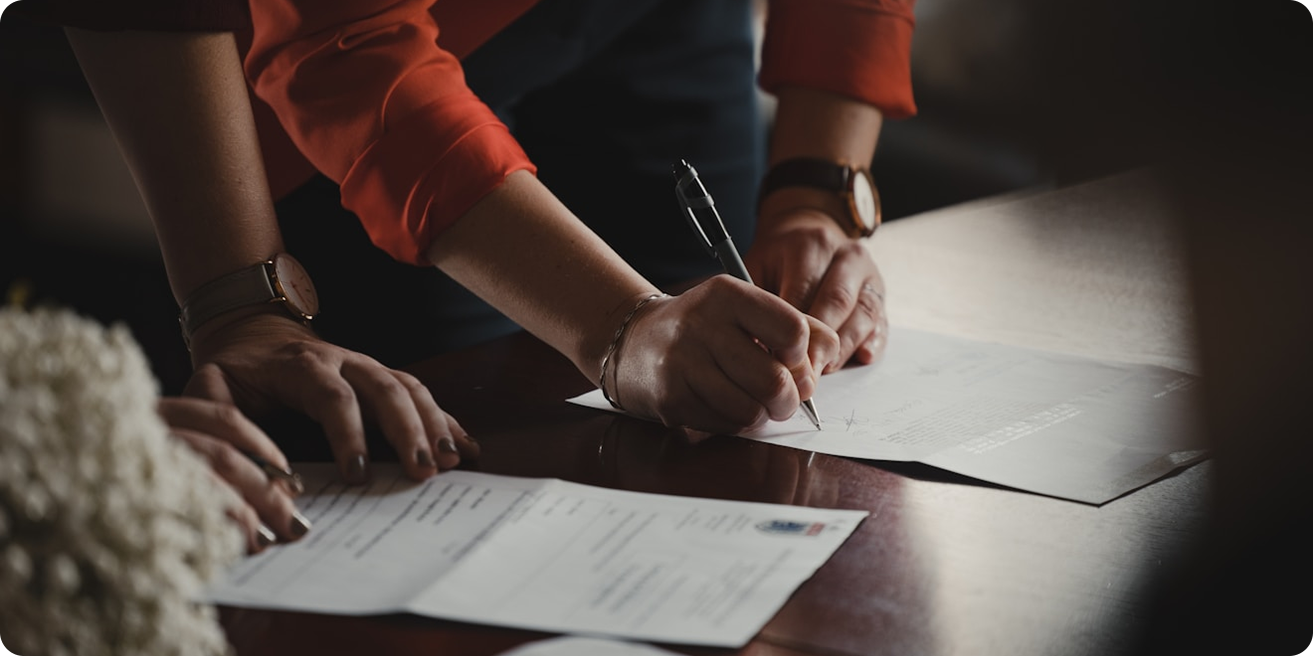 Person wearing a red long-sleeve shirt signing documents on a wooden table.