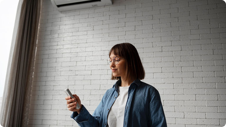Young woman in glasses and casual clothes holding and looking at a smartphone against a white brick wall.