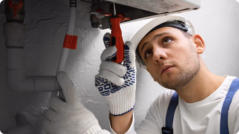 Worker wearing a white helmet and gloves using a red wrench to adjust plumbing pipes under a sink.