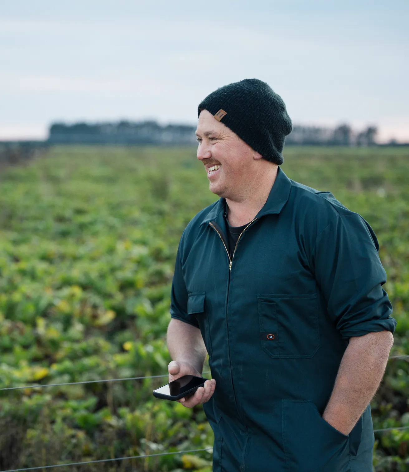 A smiling farmer wearing a dark beanie and coveralls stands in a field holding a smartphone.