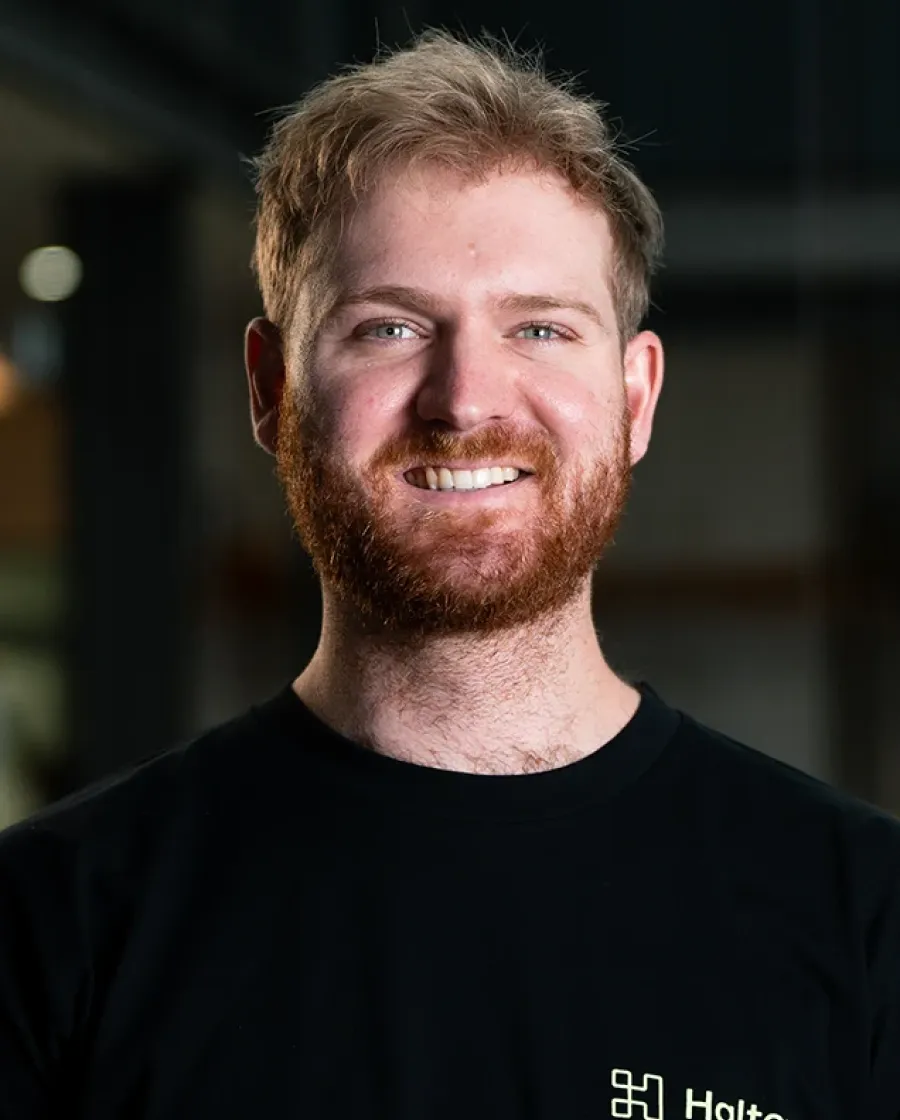 Portrait of Ben who is a smiling man with short red hair and a beard, wearing a black Halter-branded shirt, standing in a softly lit indoor setting.