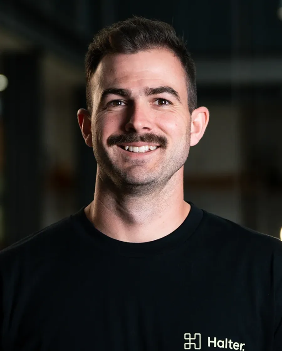 Portrait of Andy who is a smiling man with a dark mustache wearing a black Halter-branded shirt, standing indoors with blurred background lights.