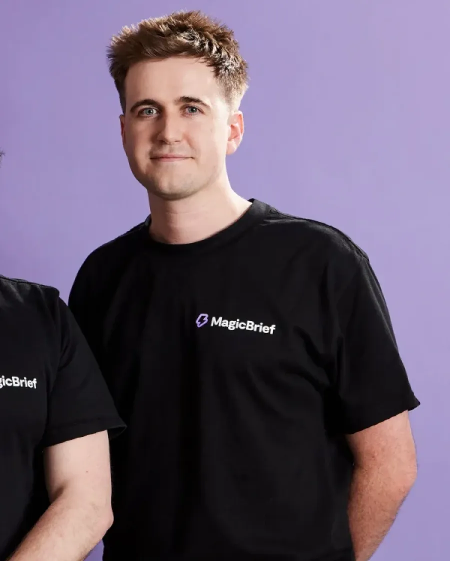 Portrait of George who is a man with short light brown hair wearing a black shirt, standing against a purple background and smiling softly.
